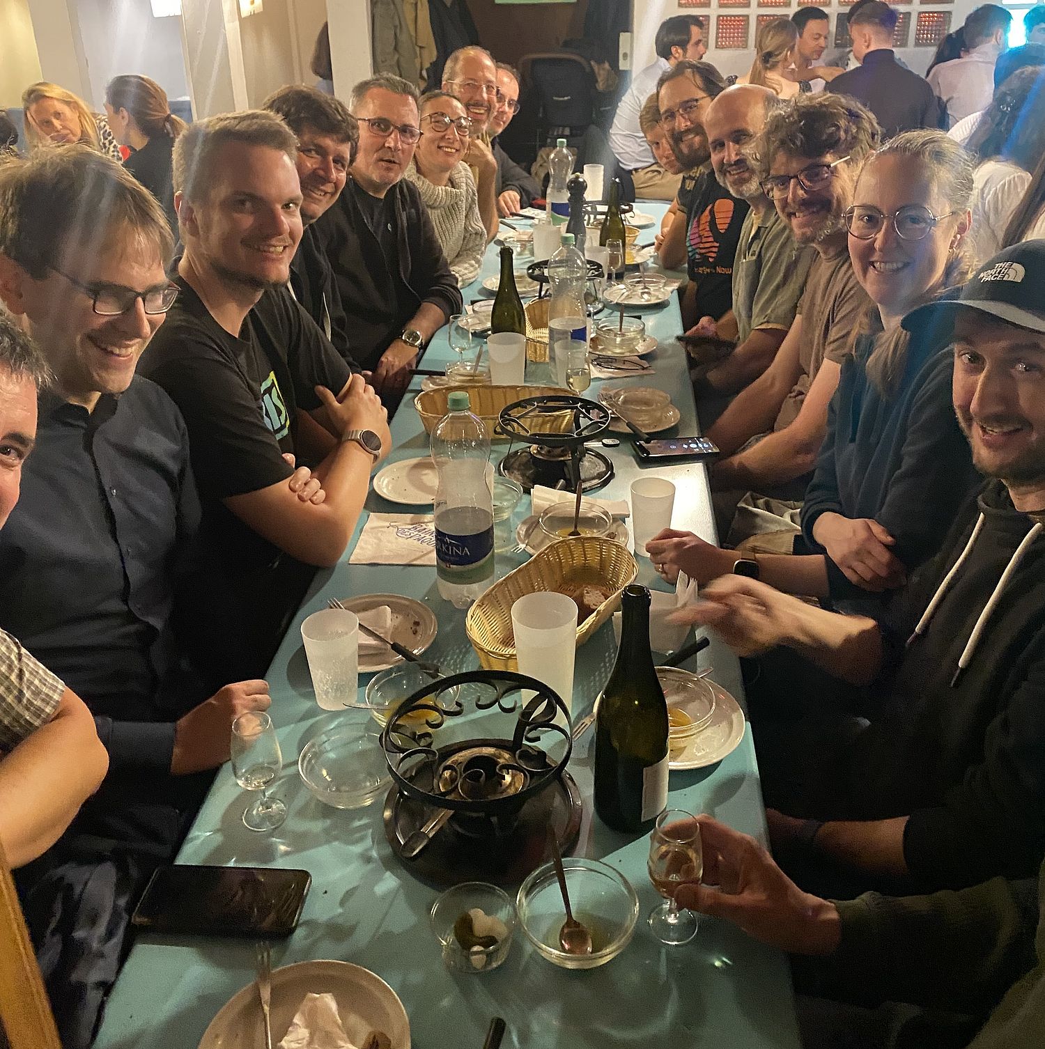 A group of people sit closely together around a long table, smiling and enjoying food and drinks at a lively indoor gathering. The table is filled with plates, bottles, and glasses.