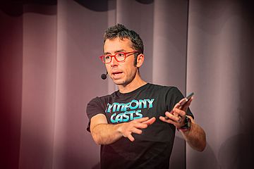 A person wearing red glasses and a black "Symfony Casts" T-shirt speaks on stage with a headset microphone, gesturing with one hand while holding a phone in the other. Gray curtains are in the background.