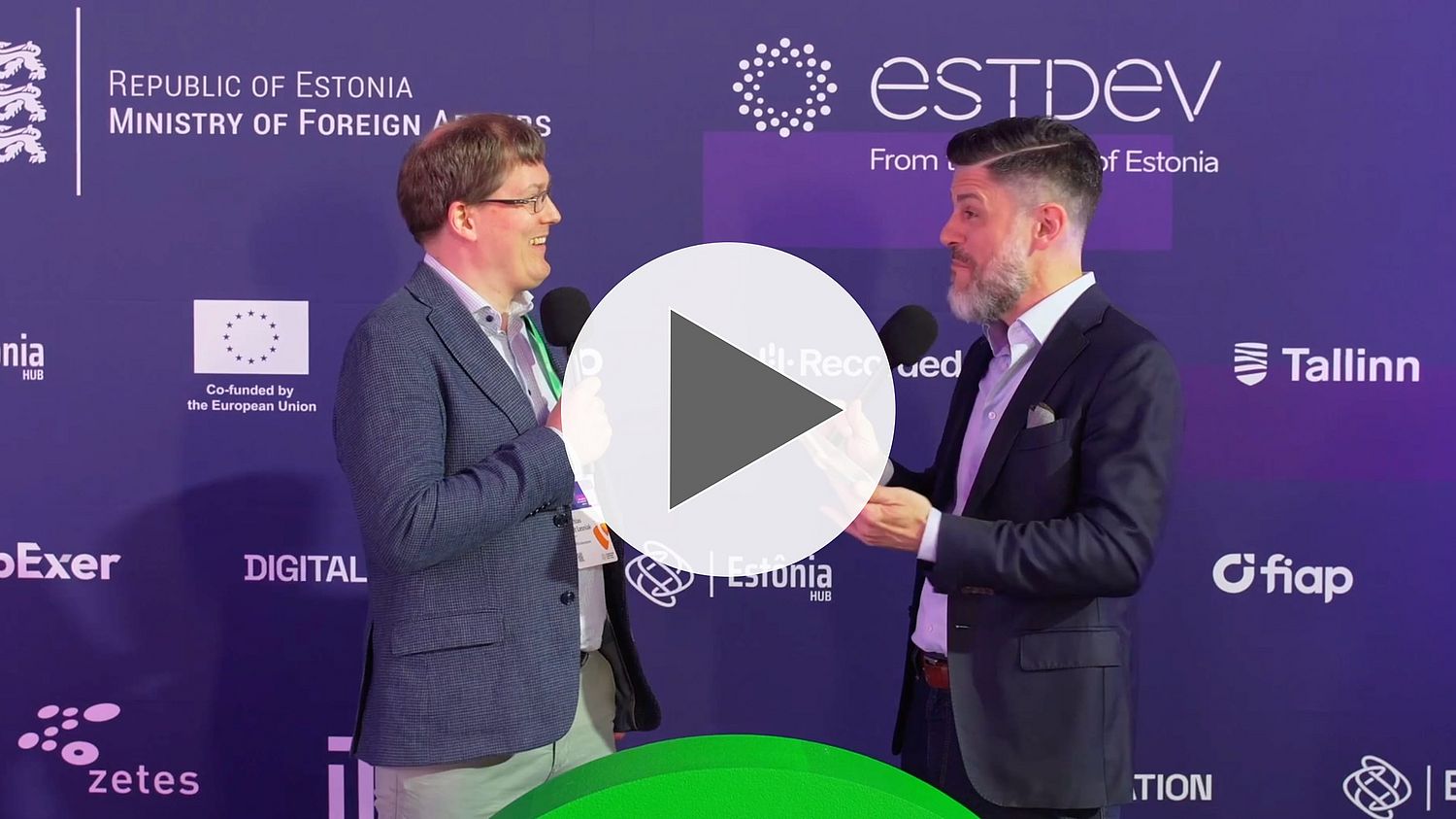 Two men in business attire stand facing each other with microphones, having a conversation in front of a purple backdrop with Estonia-related logos and text. A large play button overlays the center.