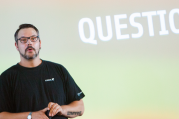 Man with beard in front of a projection of the word "questions".