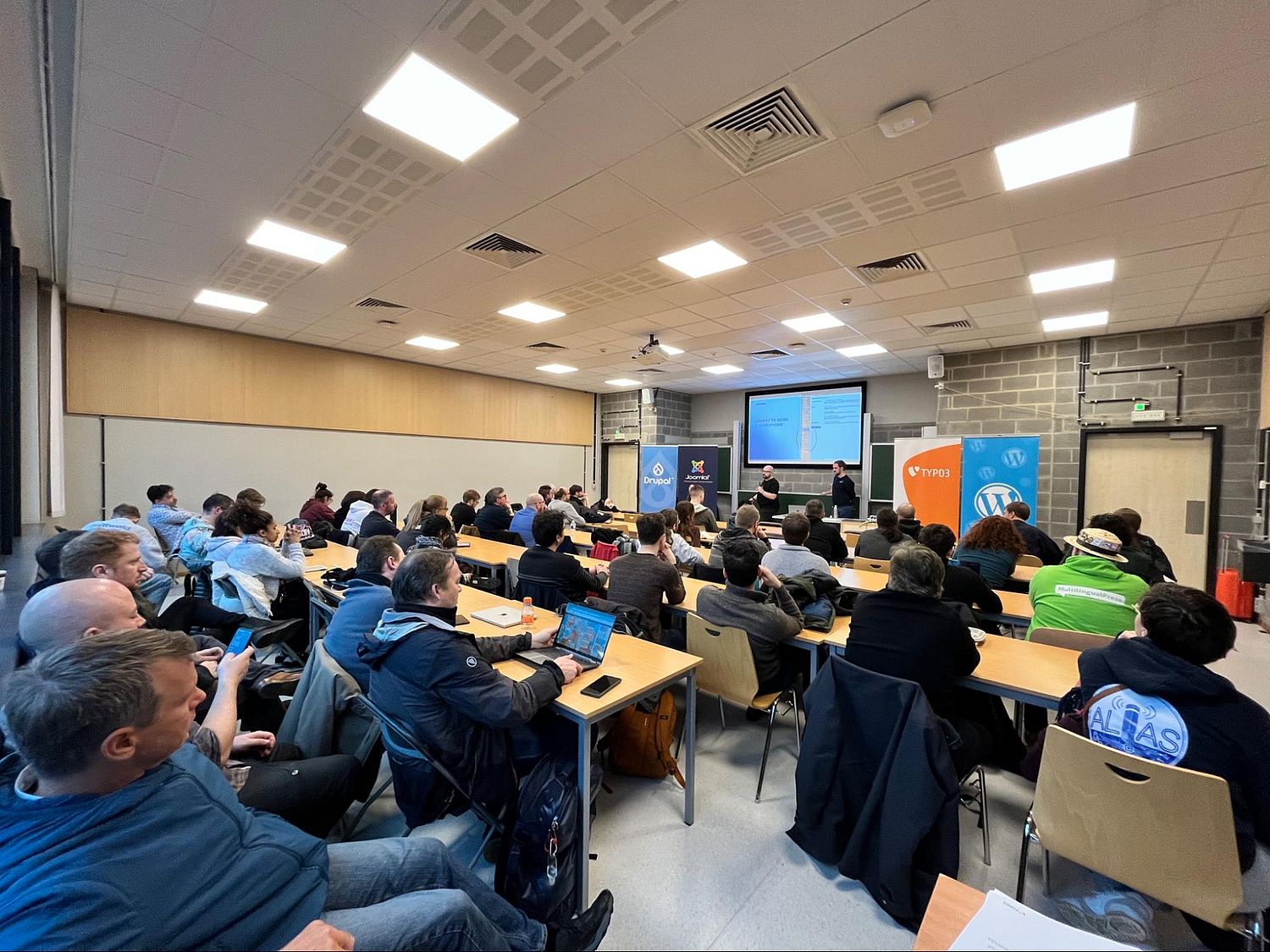 A classroom filled with people seated at desks, attentively listening to speakers at the front. Banners for Drupal, Joomla, WordPress, and TYPO3, are visible behind the speakers. The room has a projector screen displaying a presentation.