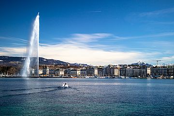 A boat sails on a large lake toward a cityscape with a tall water jet fountain and snow-capped mountains in the background under a clear blue sky.