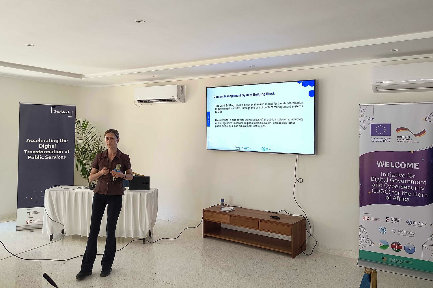 A woman stands giving a presentation in a conference room. She holds a microphone and gestures towards a screen displaying slides. Banners about digital government and cybersecurity are visible in the background.