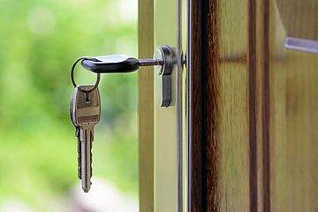 A pair of silver keys on a keyring is inserted into the lock of a wooden door, which is slightly open, with a blurred green background visible outside.