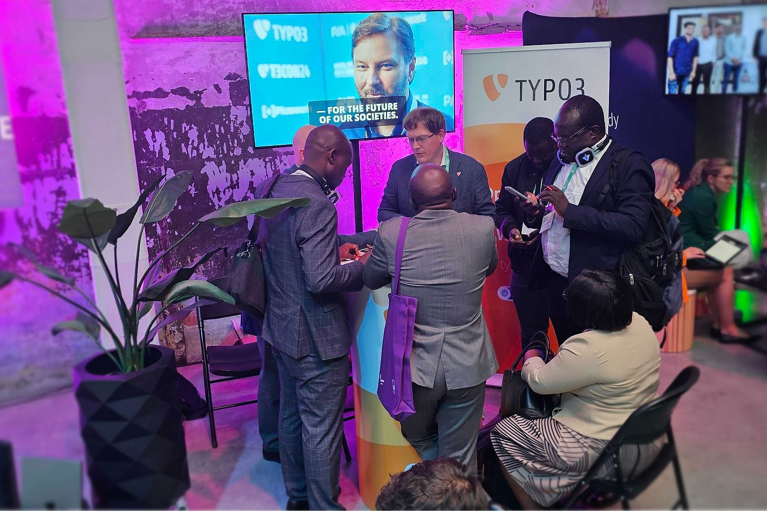 A group of people gather around a booth with TYPO3 banners at an indoor event, some standing and some sitting, engaged in conversation. A screen displays a man’s face and the text “For the future of our societies.”