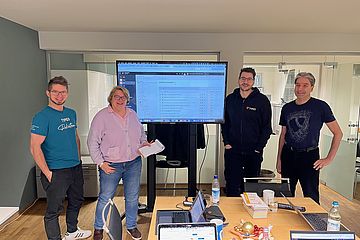 Four people stand smiling around a conference table with laptops and notebooks. A large monitor displays a webpage. The setting is a modern office with glass walls. Casual attire suggests an informal meeting.