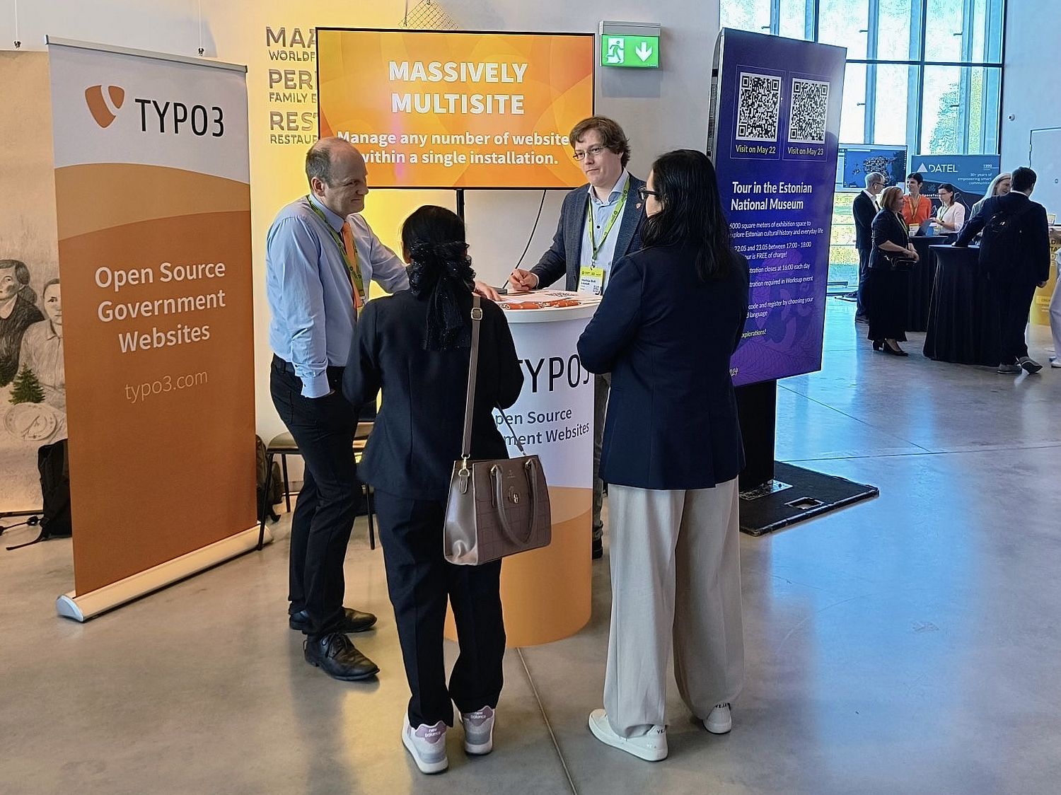 Four people in conversation at a TYPO3-branded booth with the text "massively multisite" on a display behind them.