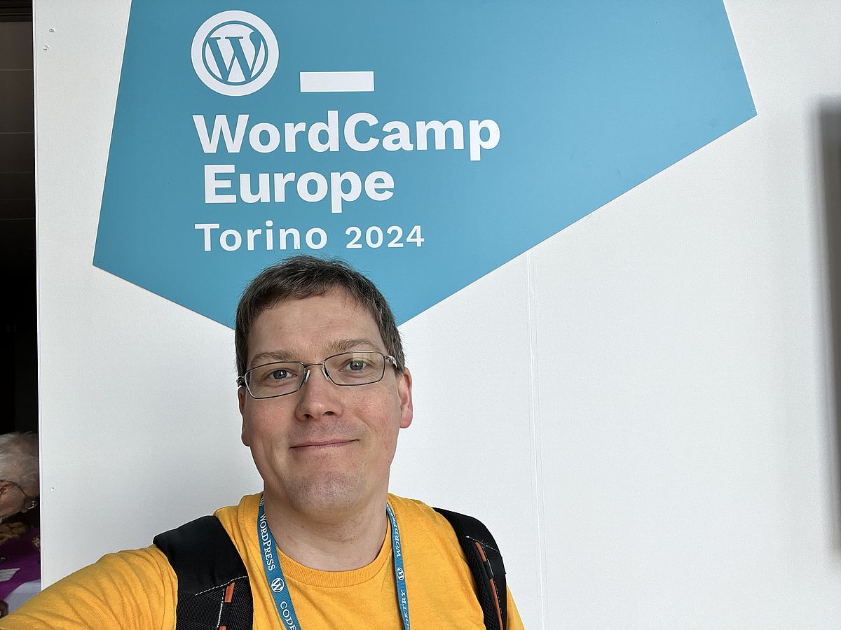 Man in yellow t-shirt in front of a white wall with a blue polygon. Within the blue polygon is the text "WordCamp Europe Torino 2024"