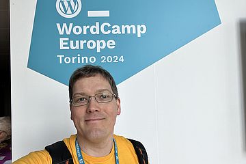 Man in yellow t-shirt in front of a white wall with a blue polygon. Within the blue polygon is the text "WordCamp Europe Torino 2024"