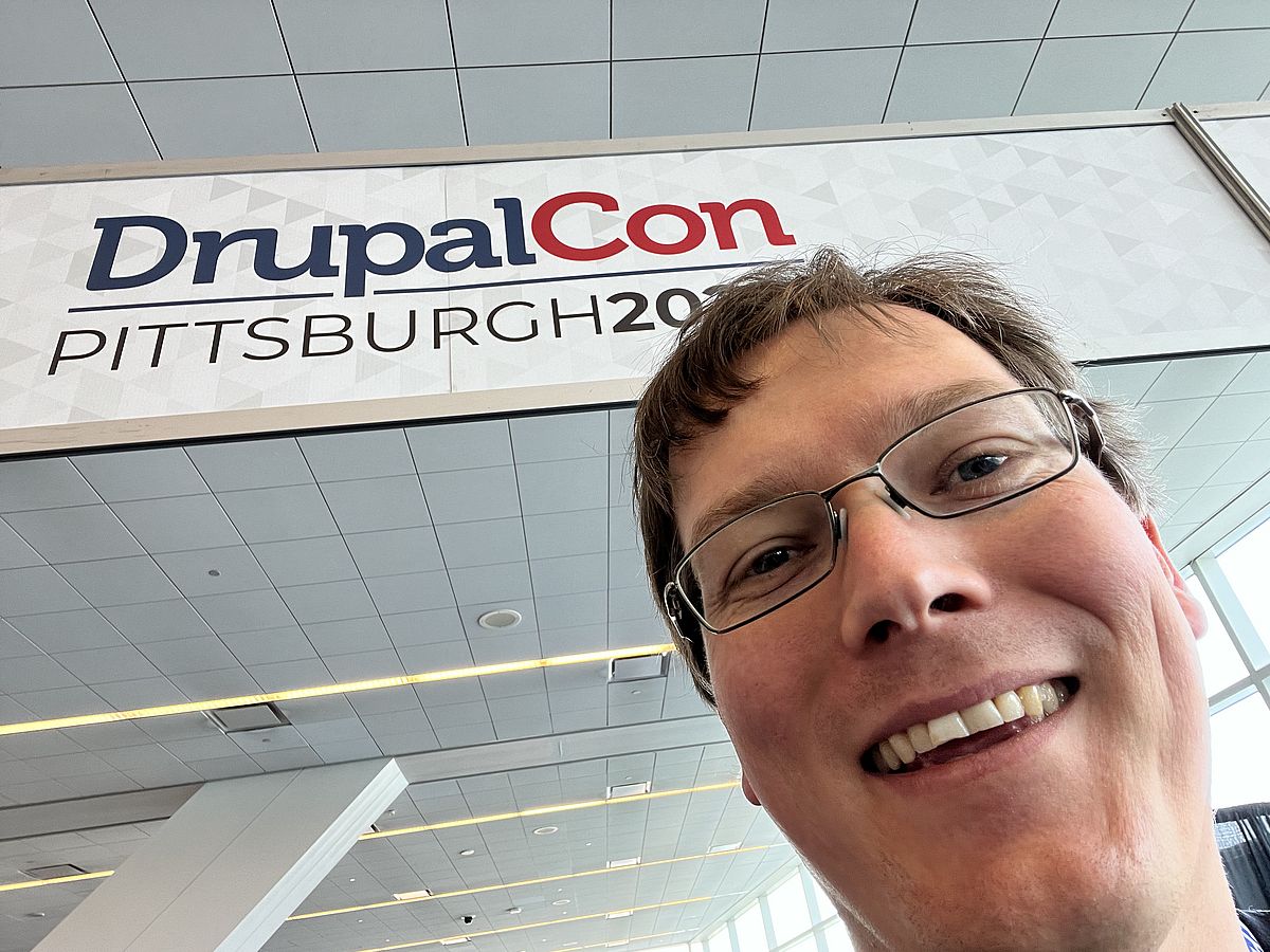Male face smiling into camera. Banner with "DrupalCon Pittsburgh 2023" in background.