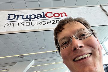 Male face smiling into camera. Banner with "DrupalCon Pittsburgh 2023" in background.