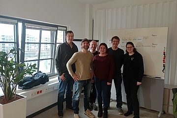 Six people standing in front of a whiteboard with a window to their right.