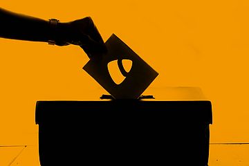 A silhouette of a hand placing a voting ballot into a box against an orange background. The ballot features a TYPO3 logo.