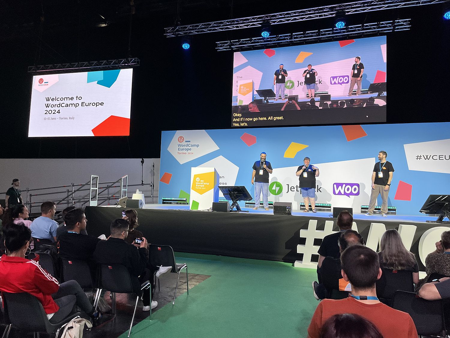 A woman and two men on a stage at a conference. A video screen above them show the three people too. A screen to the side shows the words "welcome to WordCamp Europe 2024".