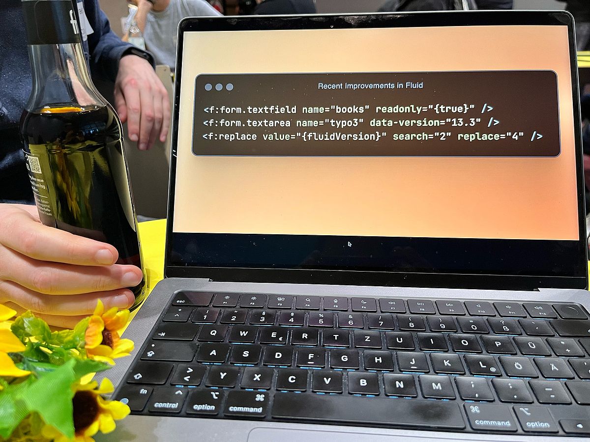 A laptop screen showing a code example. Next to it, a bottle of a black liquid and a bouquet of flowers.