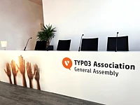 A conference panel table with four chairs and microphones, labeled TYPO3 Association General Assembly. The front features images of raised hands. A potted plant and lamp are in the background.