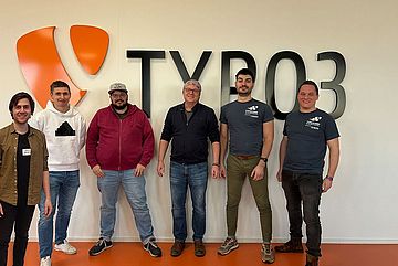 Six people stand smiling in front of a wall with a large TYPO3 logo and text. The group consists of five men and one woman, all casually dressed, in a brightly lit room with an orange floor.