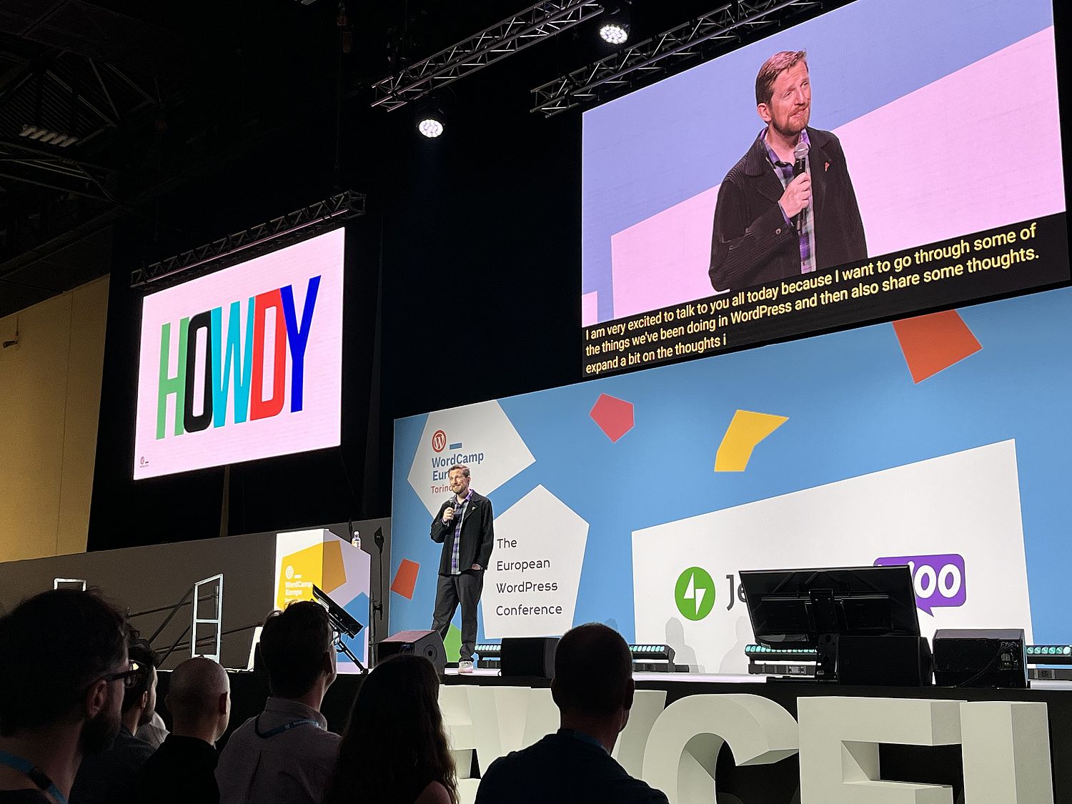 A single man on stage with the word "howdy" in multicolored uppercase letters on a projection screen in the background.