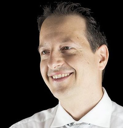 A man in a white dress shirt and light-colored tie smiles and looks to the side with his arms crossed, standing against a black background.