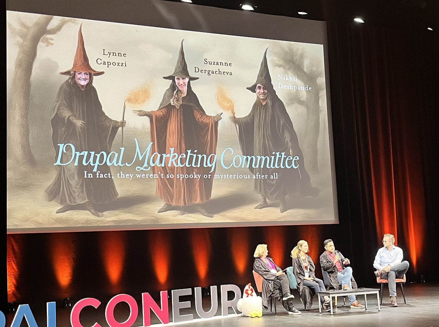 Stage with four people seated below a projection screen showing three persons dressed as witches or wizards. Label: Drupal Marketing Committee.