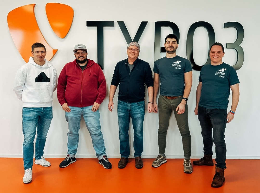 Five men in casual clothes standing on an orange floor. Behind them, the TYPO3 logo on a white wall.