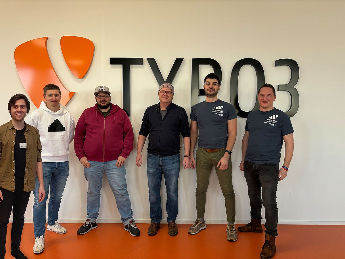 Six people stand smiling in front of a wall with a large TYPO3 logo and text. The group consists of five men and one woman, all casually dressed, in a brightly lit room with an orange floor.